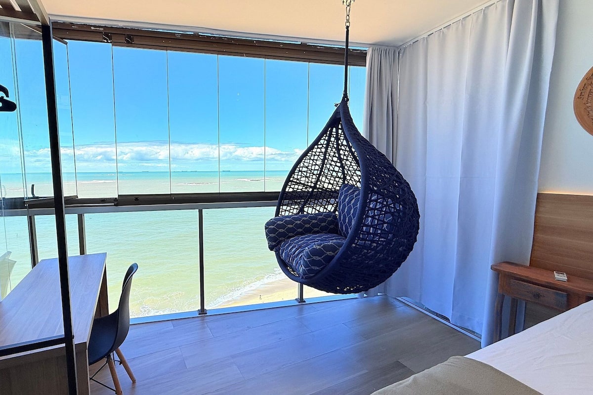 Una habitación con un ventanal grande con vista a una playa, una silla colgante, una mesa de madera y una cama.