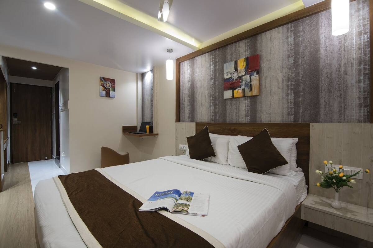 A hotel room with a large bed, a bedside table with a vase of flowers, a wall-mounted artwork, and a door leading to another room.