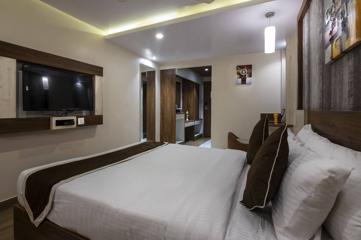 A modern hotel room with a large bed, a flat-screen TV mounted on the wall, a desk with a laptop, and a small painting on the wall.