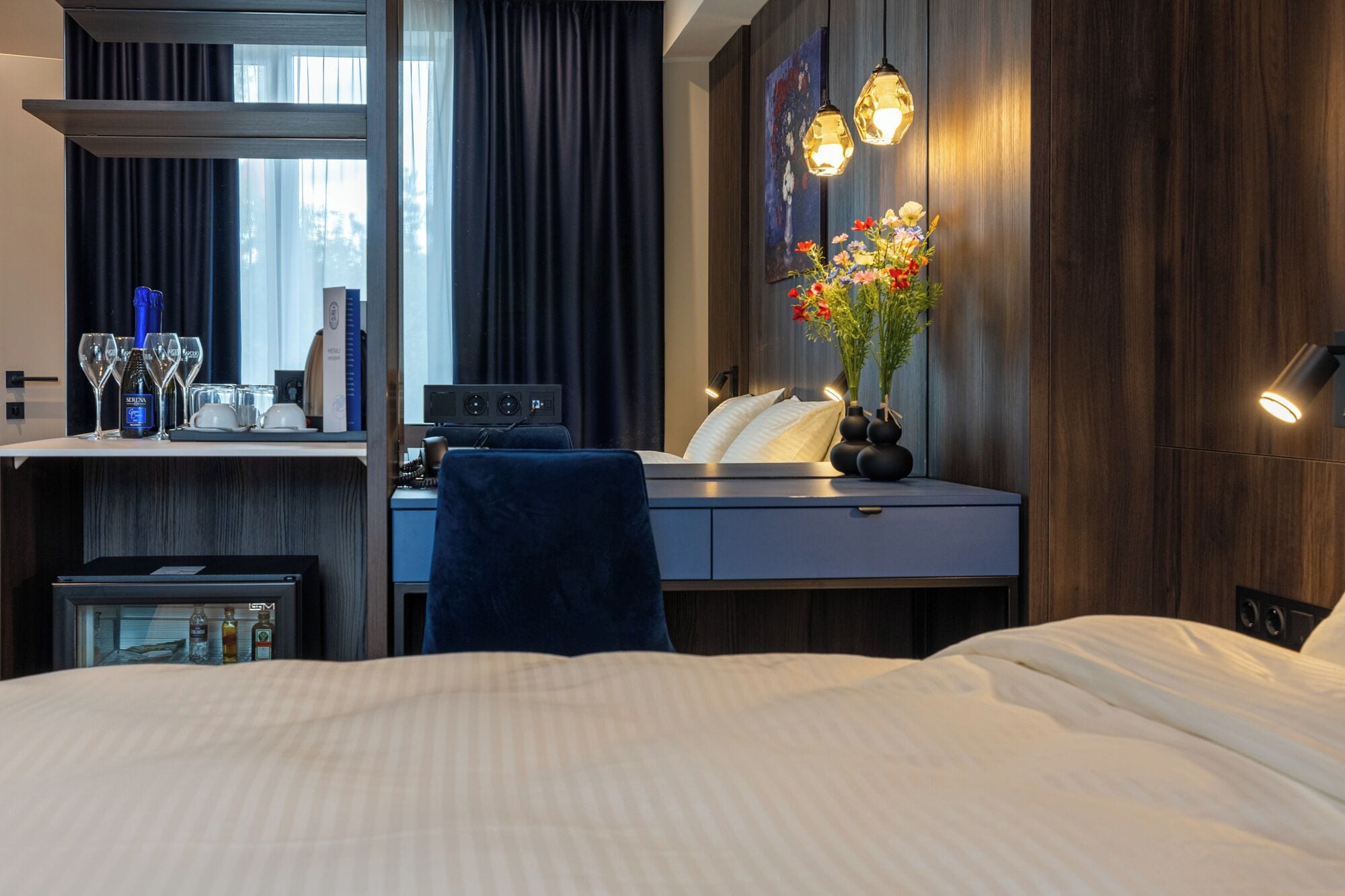 A hotel room with a bed, a desk with a computer, a chair, a small table with a bottle and glasses, a vase with flowers, and a wall-mounted light fixture.