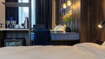 Ein Hotelzimmer mit einem Bett, einem Schreibtisch mit Computer, einem Sessel, einem kleinen Tisch mit Flasche und Gläsern, einer Vase mit Blumen und einer Wandleuchte.