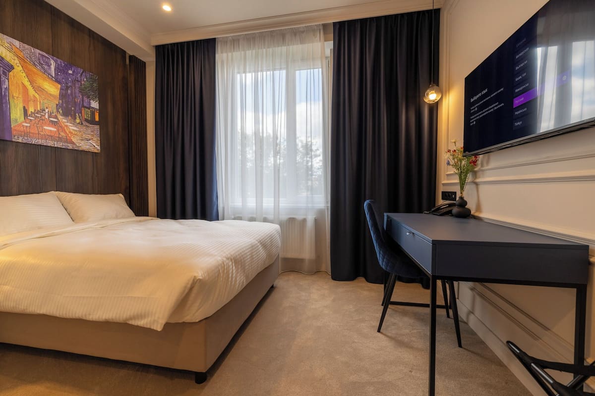A hotel room with a bed, a desk, a chair, a TV, and a painting on the wall.