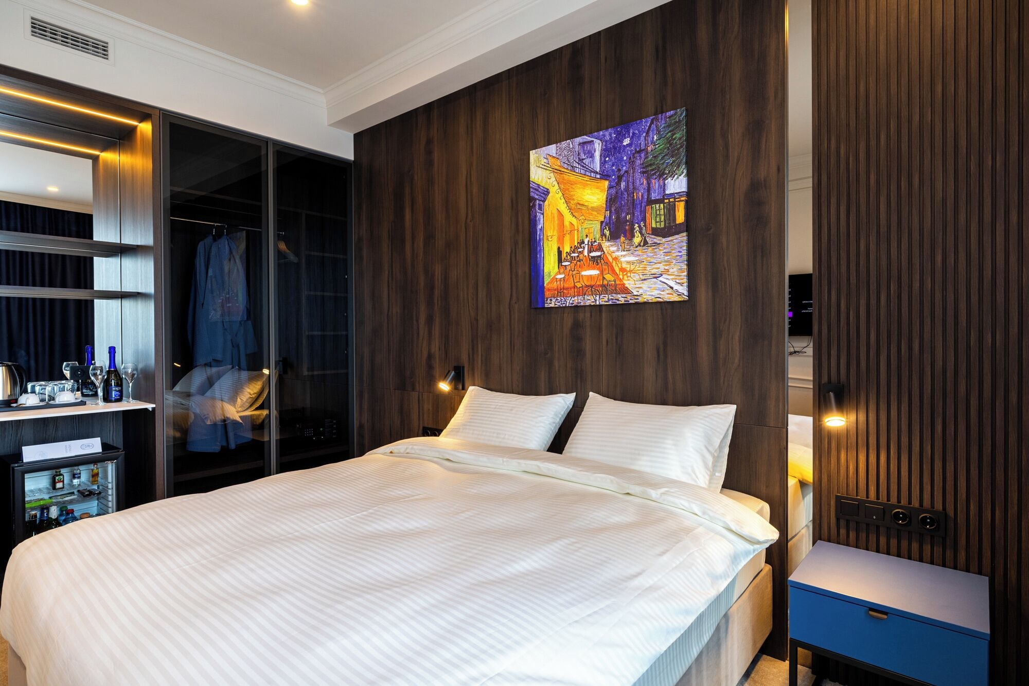 A modern hotel room with a large bed, a wardrobe with a mirror, a painting on the wall, and a small table with a lamp.