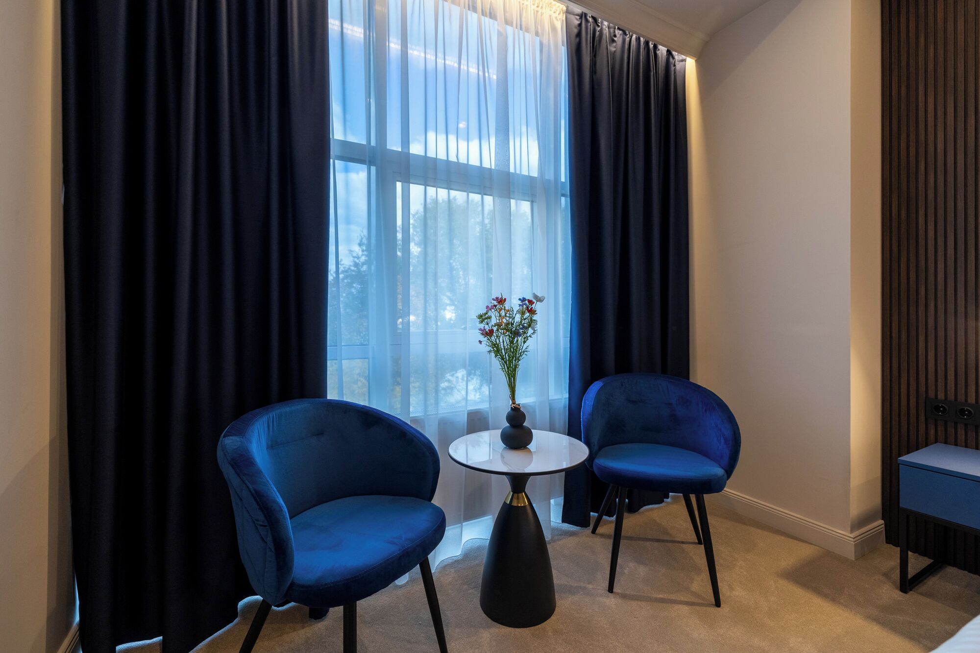 A room with two blue armchairs, a small round table, a vase with flowers, and a view through sheer curtains.