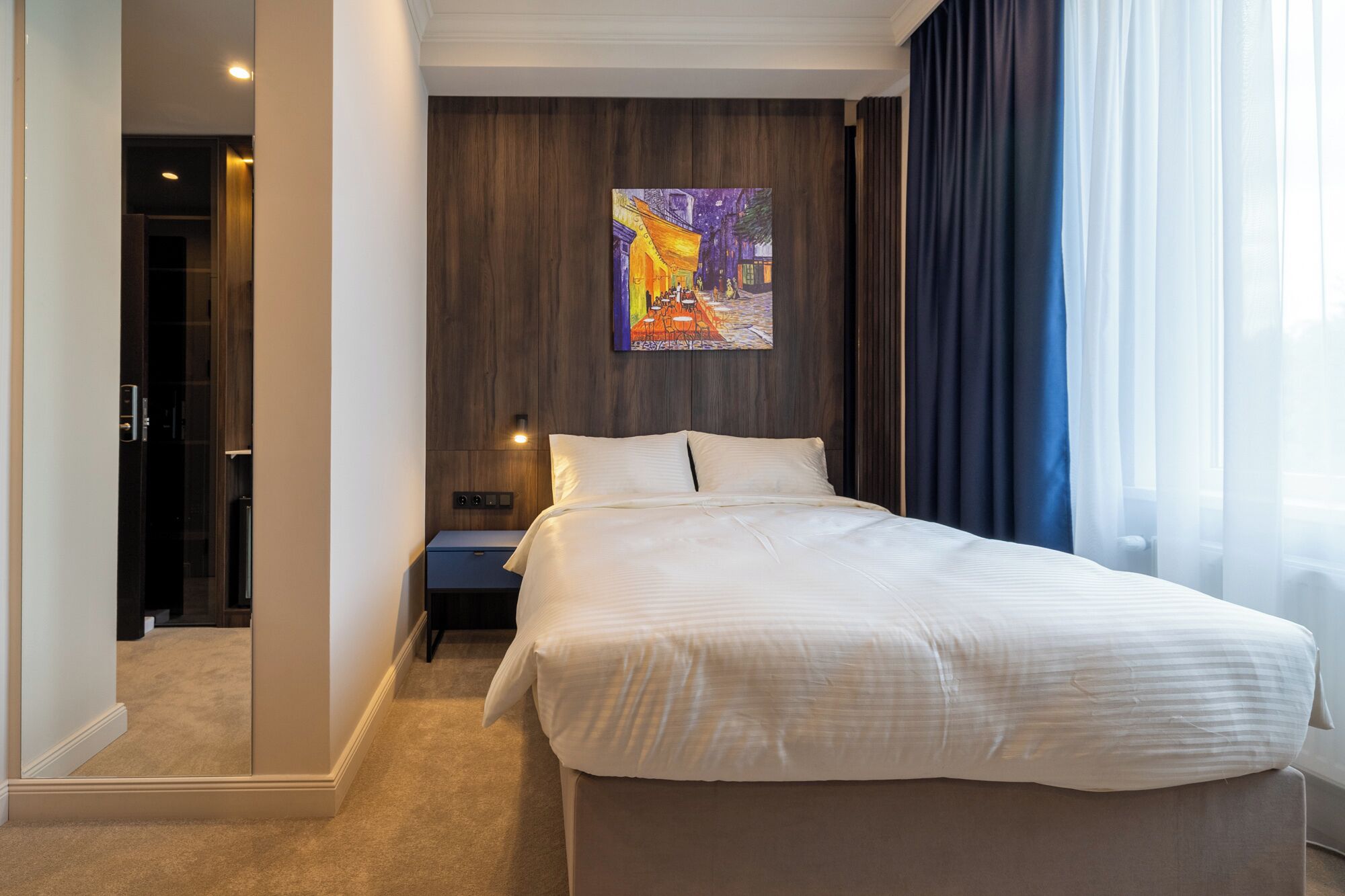 Una camera d'albergo con un letto grande, un comodino, un quadro appeso e vista sull'esterno attraverso tende leggere.
