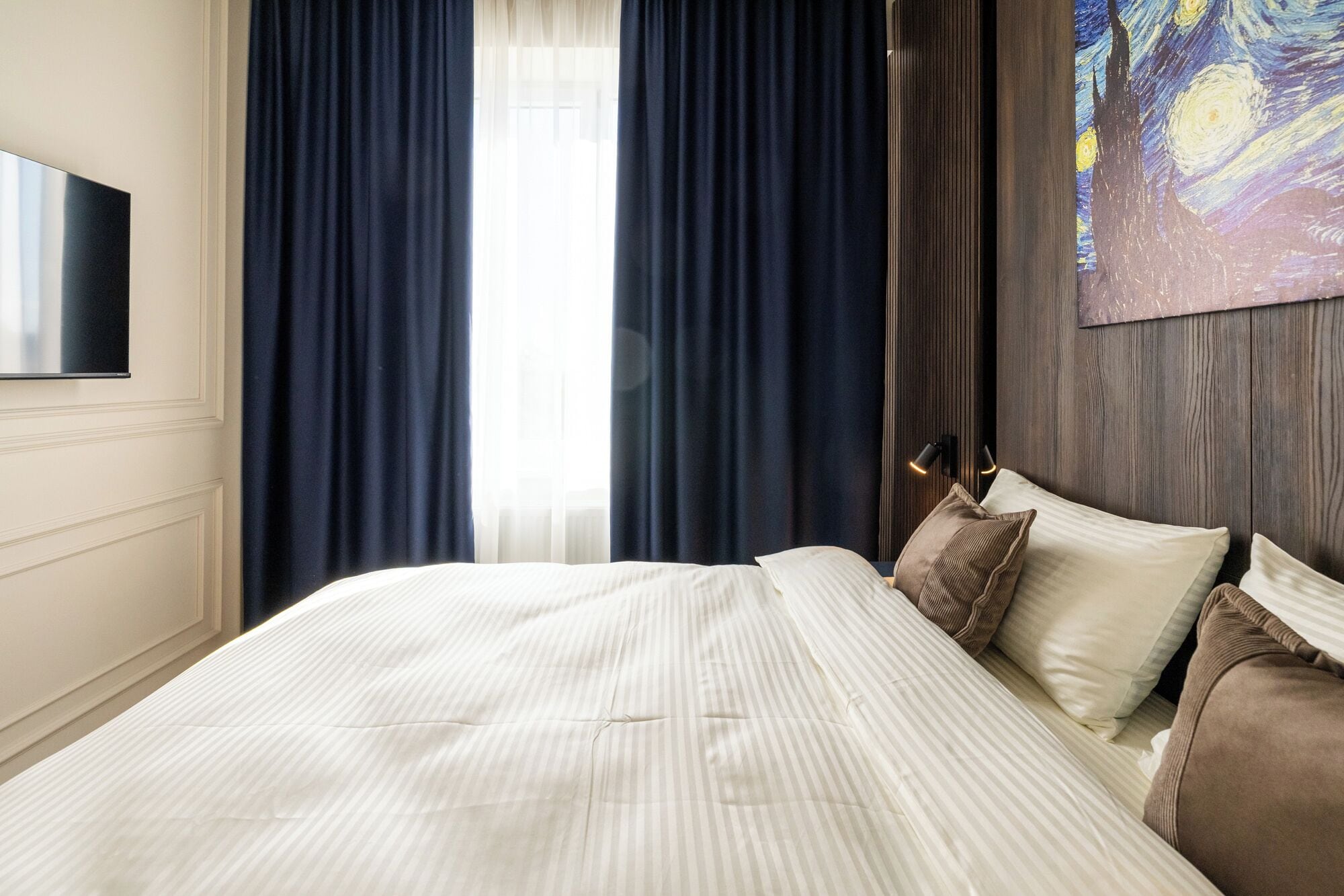 A hotel room with a large bed, dark curtains, a painting on the wall, and a flat-screen TV.