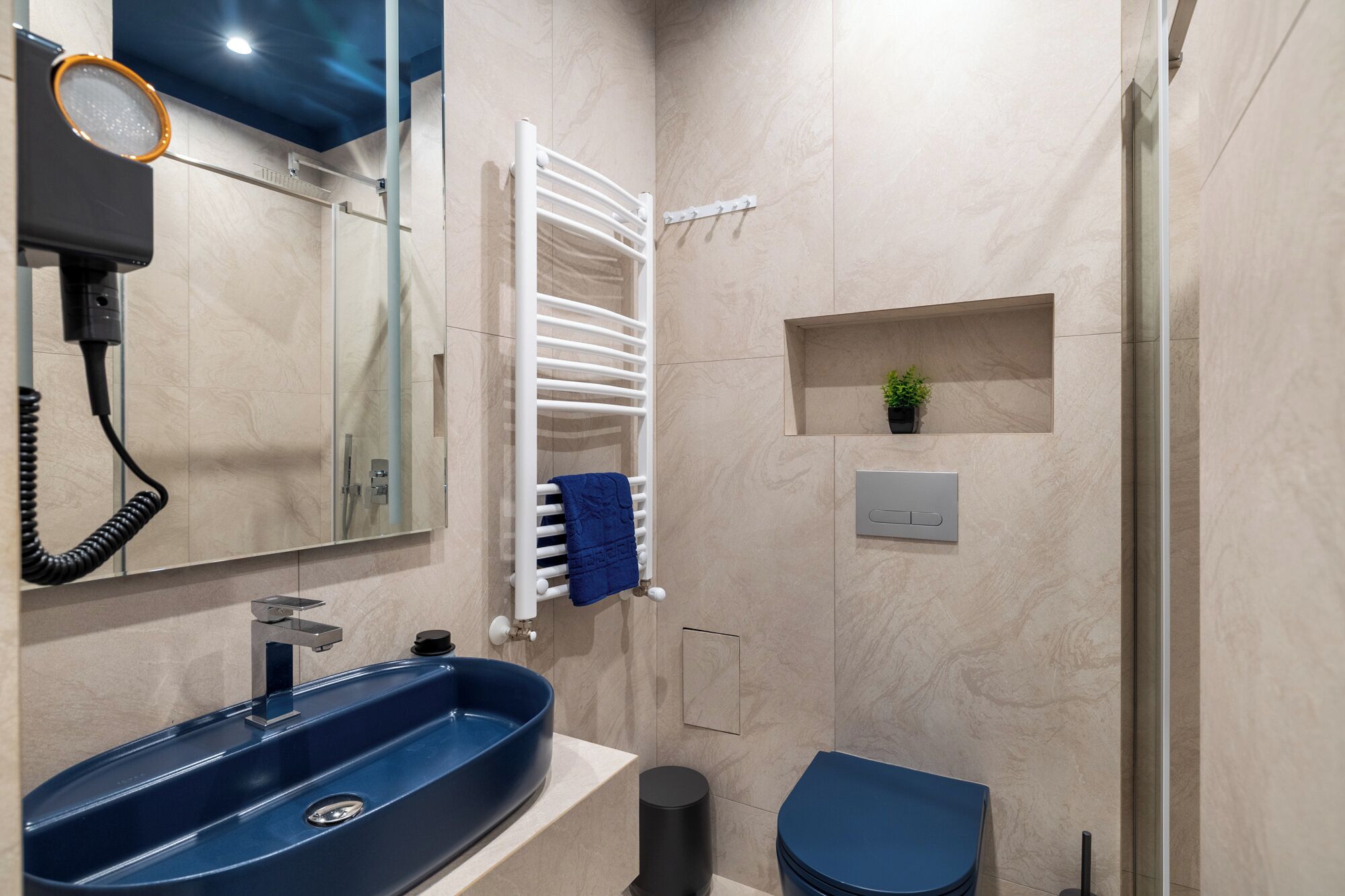 A modern bathroom with a blue sink, a white towel rack, a small plant, and a toilet.