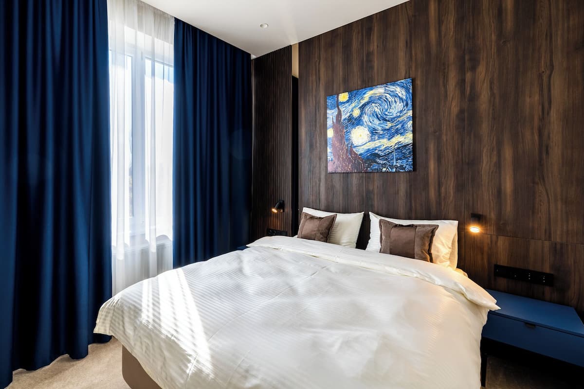 A modern hotel room with a large bed, dark wooden paneling, a painting on the wall, and a view of the city through the window.