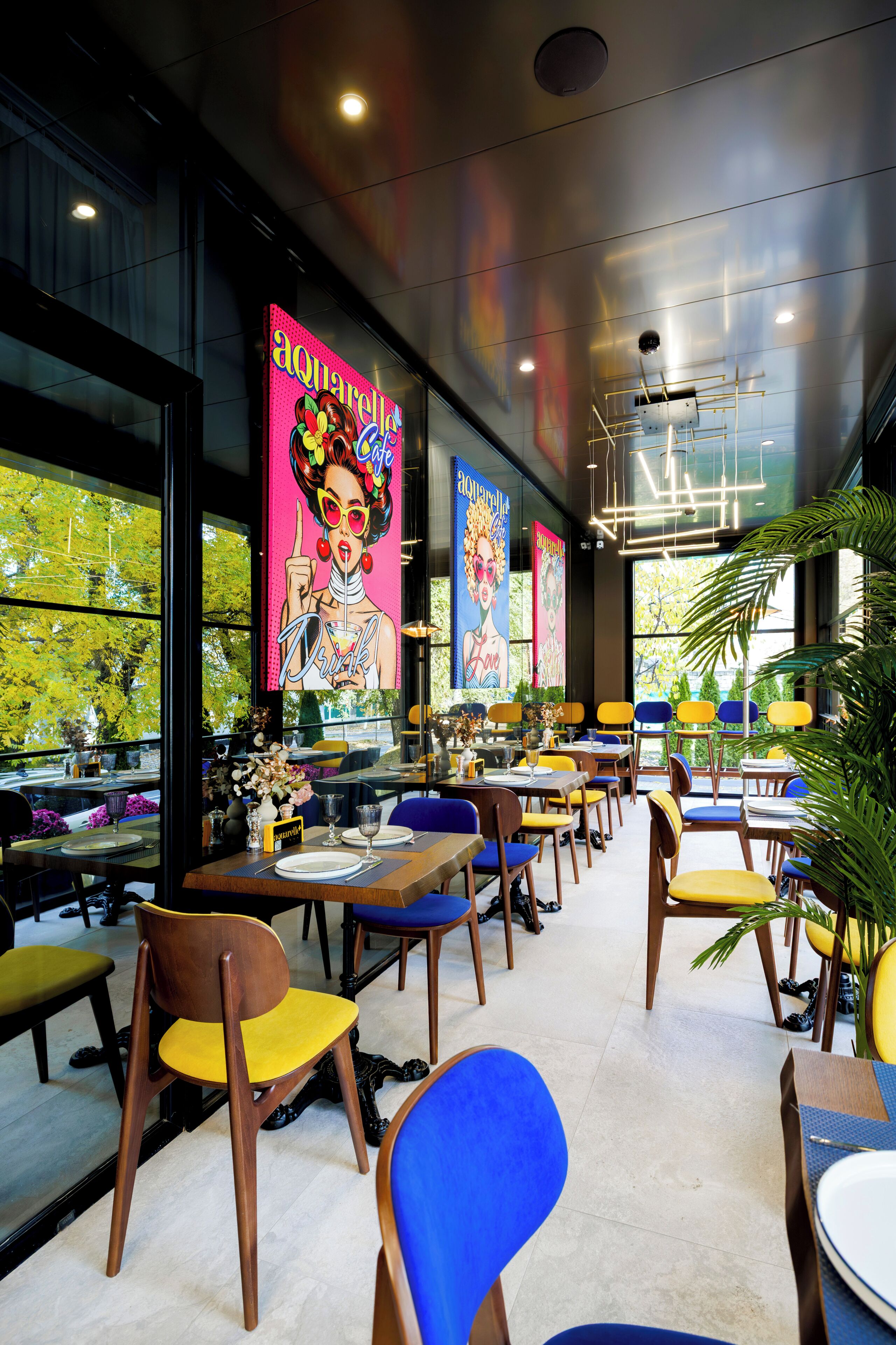 A restaurant with multiple tables and chairs, large windows, and a ceiling with exposed lighting fixtures.