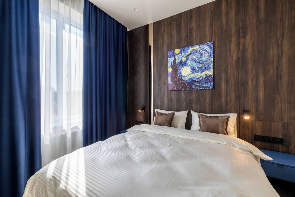 A modern hotel room with a large bed, dark wooden paneling, a painting on the wall, and a view of the city.