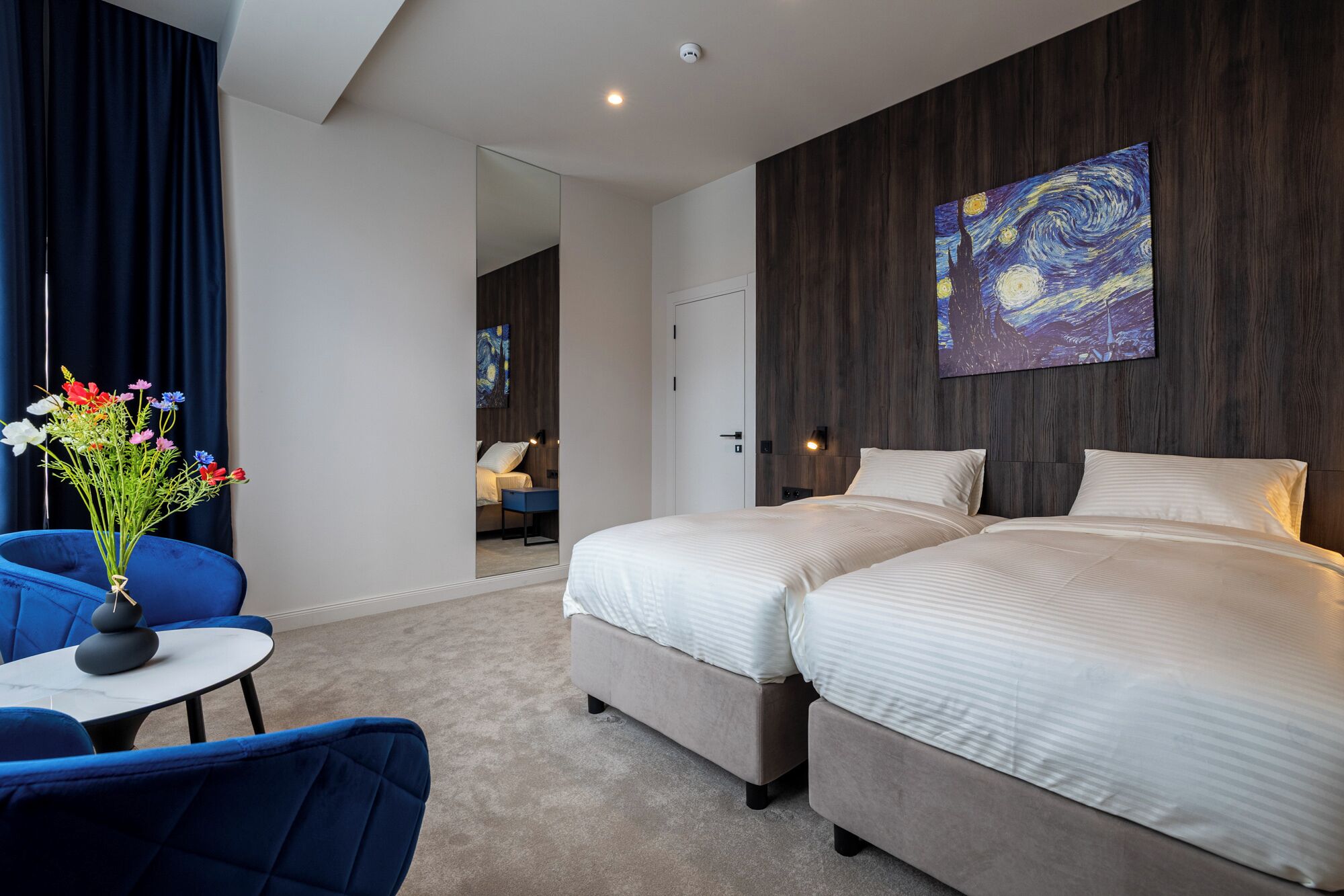Una habitació d'hotel moderna amb dos llits, una cadira blava, una taula rodona petita i un quadre a la paret.