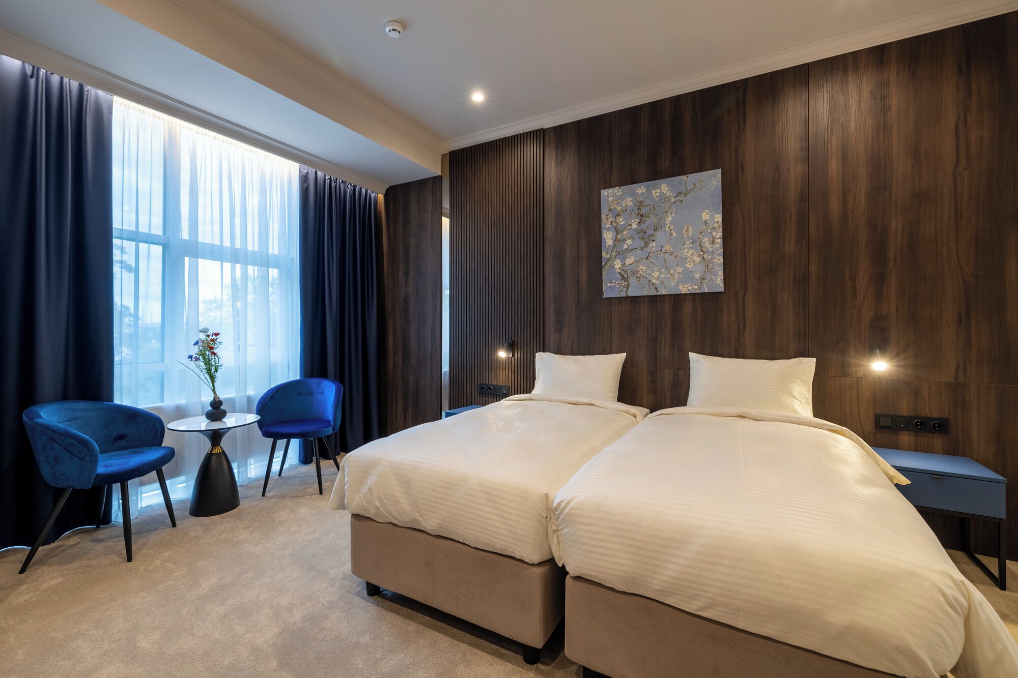A hotel room with a large bed, a small table with a vase, two blue chairs, and a large window with curtains.