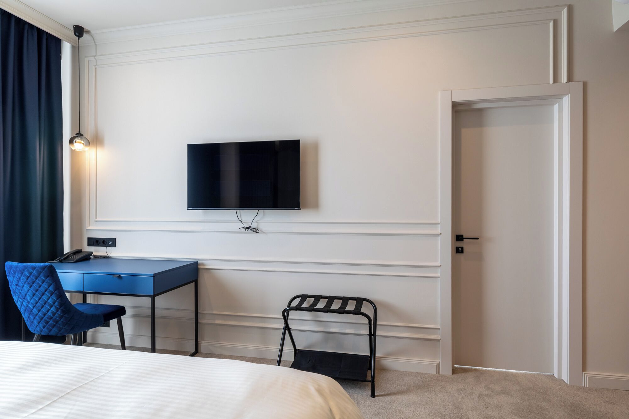 Una habitación de hotel con cama, escritorio con silla, televisor montado en la pared y una puerta.