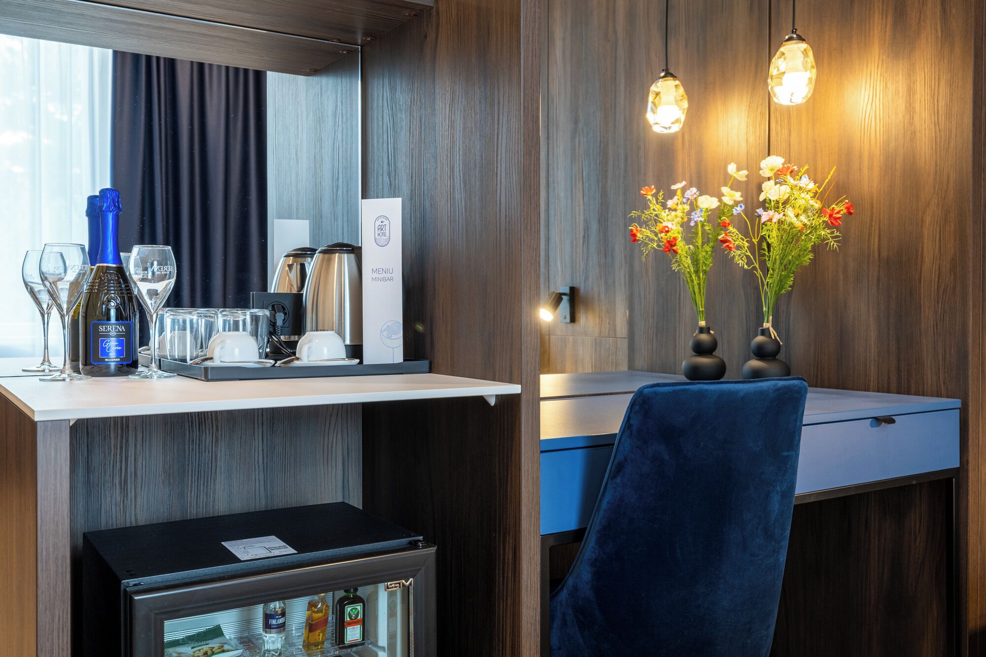 Hotellihuone, jossa on minibaari, minibaarin jääkaappi, työpöytä tuolilla sekä seinään kiinnitetty valaisin.