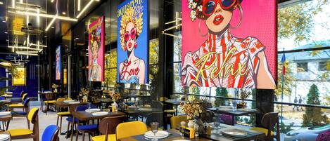 A modern restaurant interior with tables, chairs, and large colorful posters on the walls.
