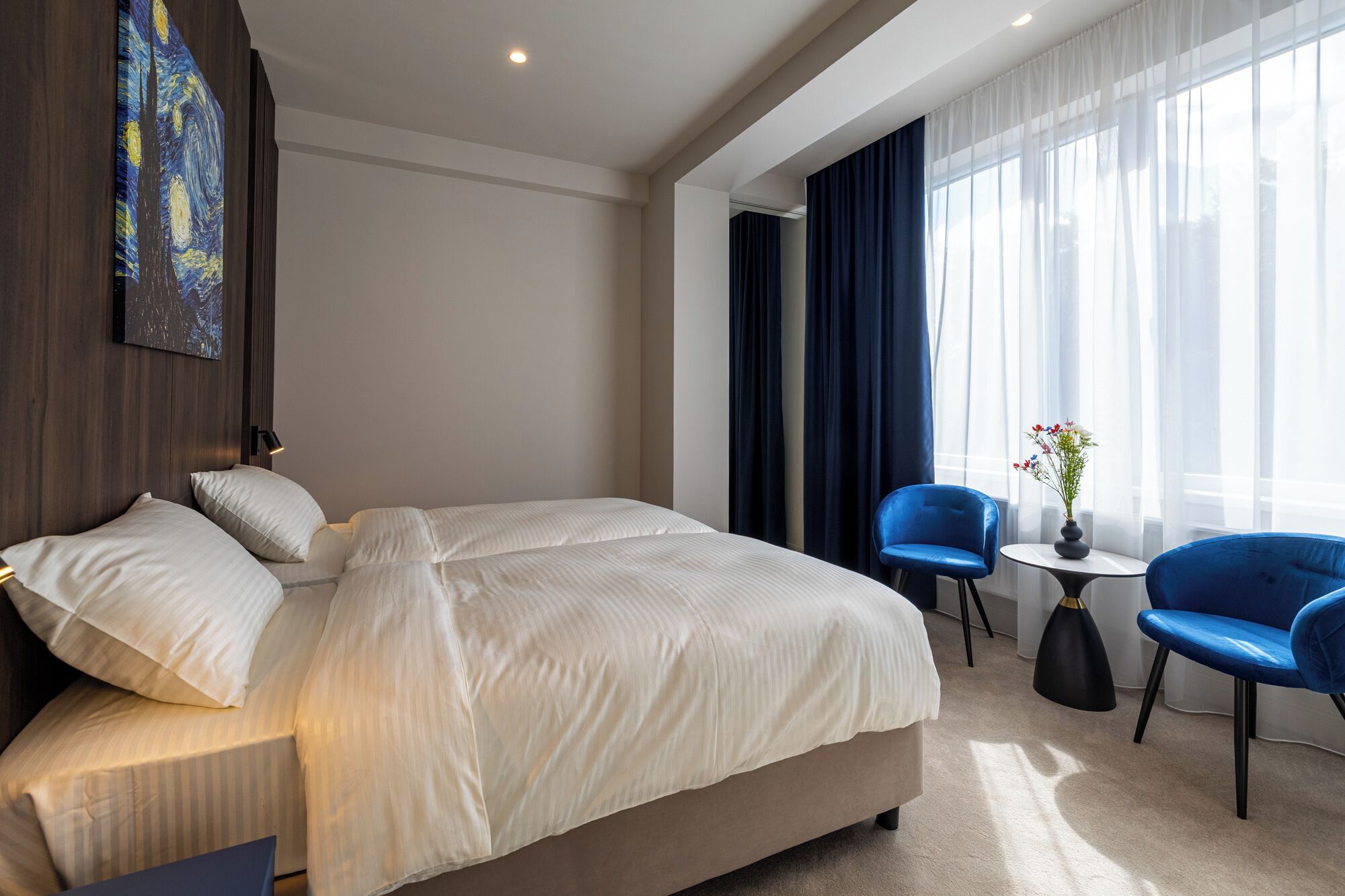 Quarto de hotel moderno com uma cama grande, duas cadeiras azuis, uma mesinha com um vaso e um quadro na parede.
