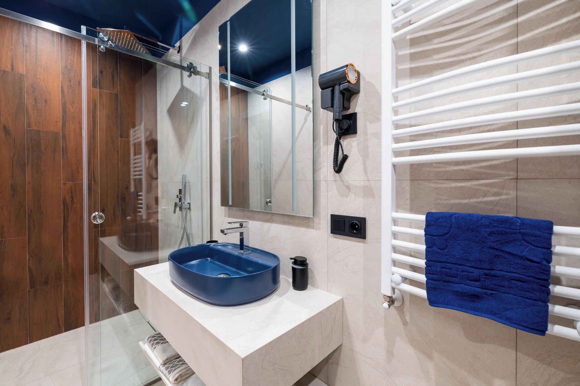 A modern bathroom with a glass-enclosed shower, a blue sink, a mirror, a towel rack, and a hairdryer.