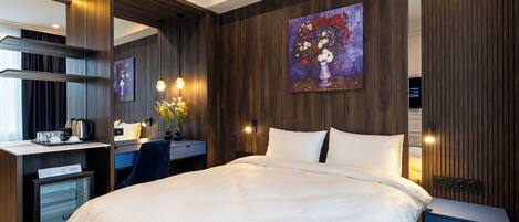 A modern hotel room with a large bed, a desk with a kettle, and a minibar.