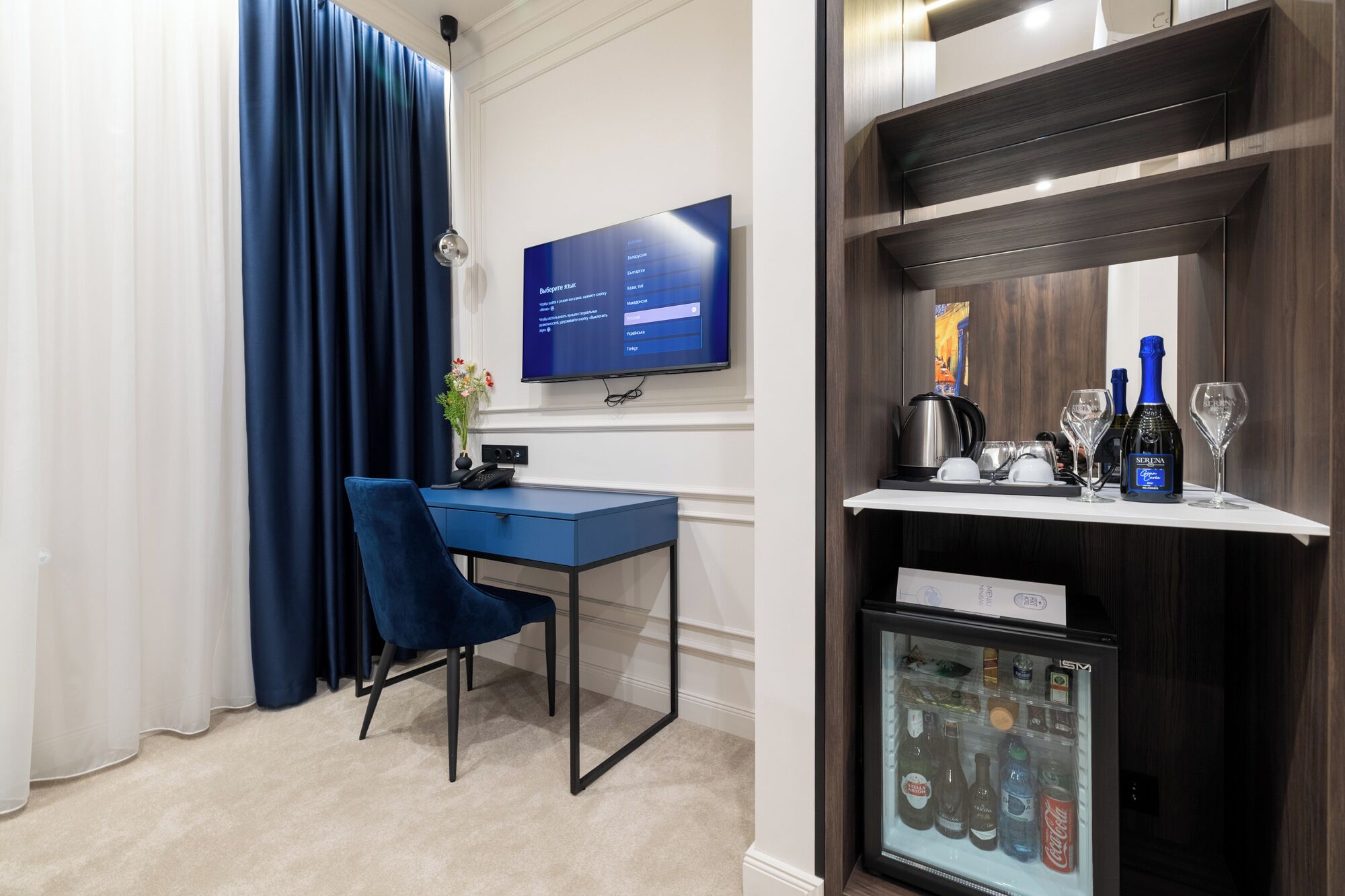 A hotel room with a desk, chair, TV, minibar, and a coffee machine.