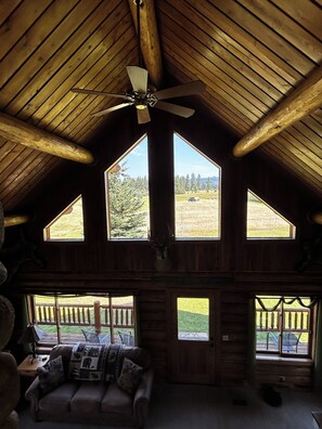 A cabin interior with a high ceiling, a ceiling fan, and large windows offering a view of a grassy landscape. - Amazing mountain views, daily wildlife show and a cozy log house for serenity. (Seeley Lake)