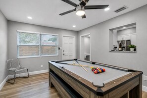 A pool table with billiard balls and cues, a ceiling fan, and a window with blinds.