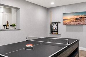 A ping pong table with a net, a pool table with balls, a rack of billiard balls, a wine bottle, glasses, and a small plant.
