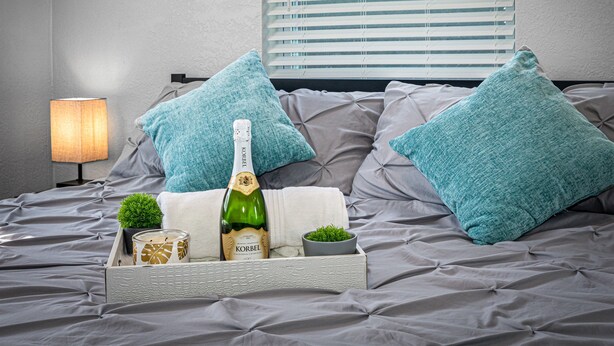 A bed with grey bedding, two teal pillows, a bottle of Korbel champagne, a small plant, and a candle on a tray. - Heated Pool & Game Room, Sleeps 10 | 5 Min to Busch Gardens, Tampa (Tampa)