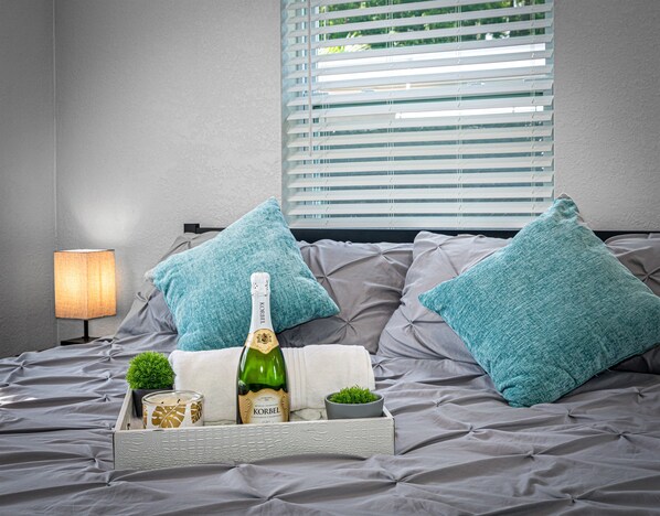 Una cama con ropa de cama gris, dos almohadas color turquesa, una botella de champán Korbel, una planta pequeña y una vela sobre una bandeja.