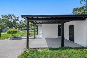 Un edificio moderno con un patio techado, un camino de hormigón y una pequeña zona ajardinada con un arbusto.