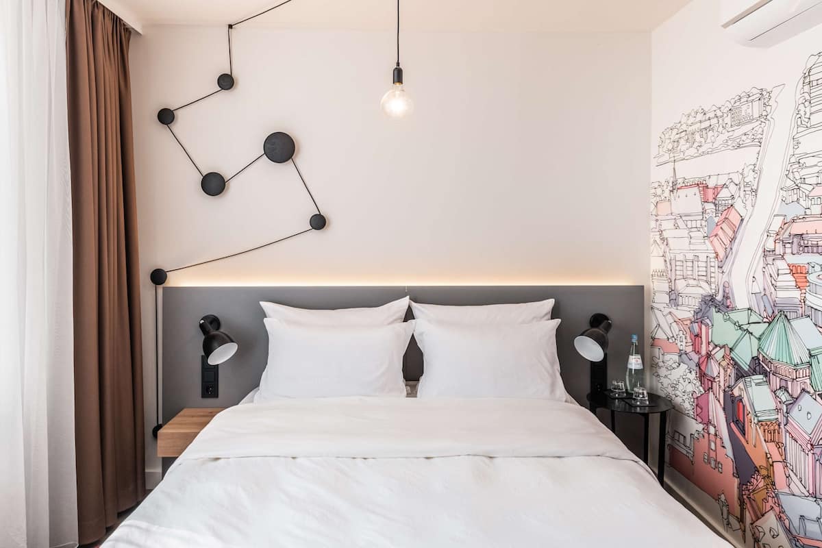 A modern hotel room with a neatly made bed, a wall-mounted light fixture, and a colorful cityscape mural.