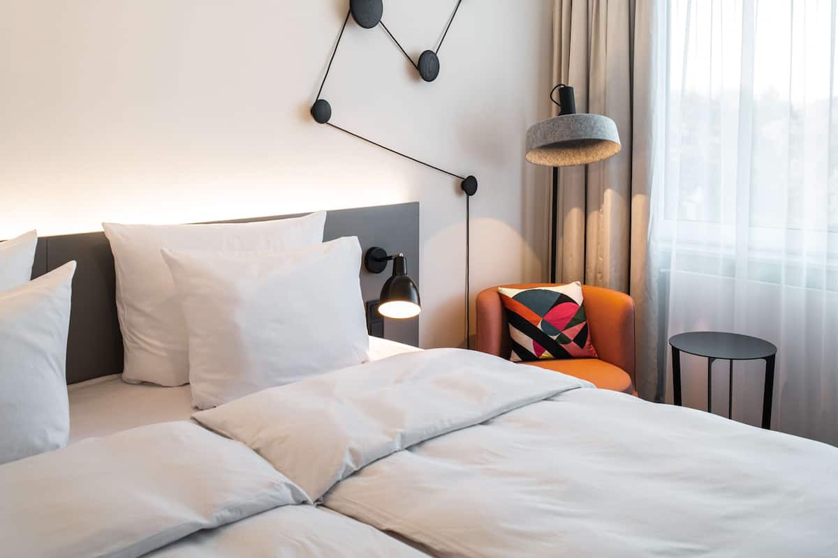 A neatly made bed with white linens, a modern wall-mounted lamp, and a colorful chair by a window with sheer curtains.