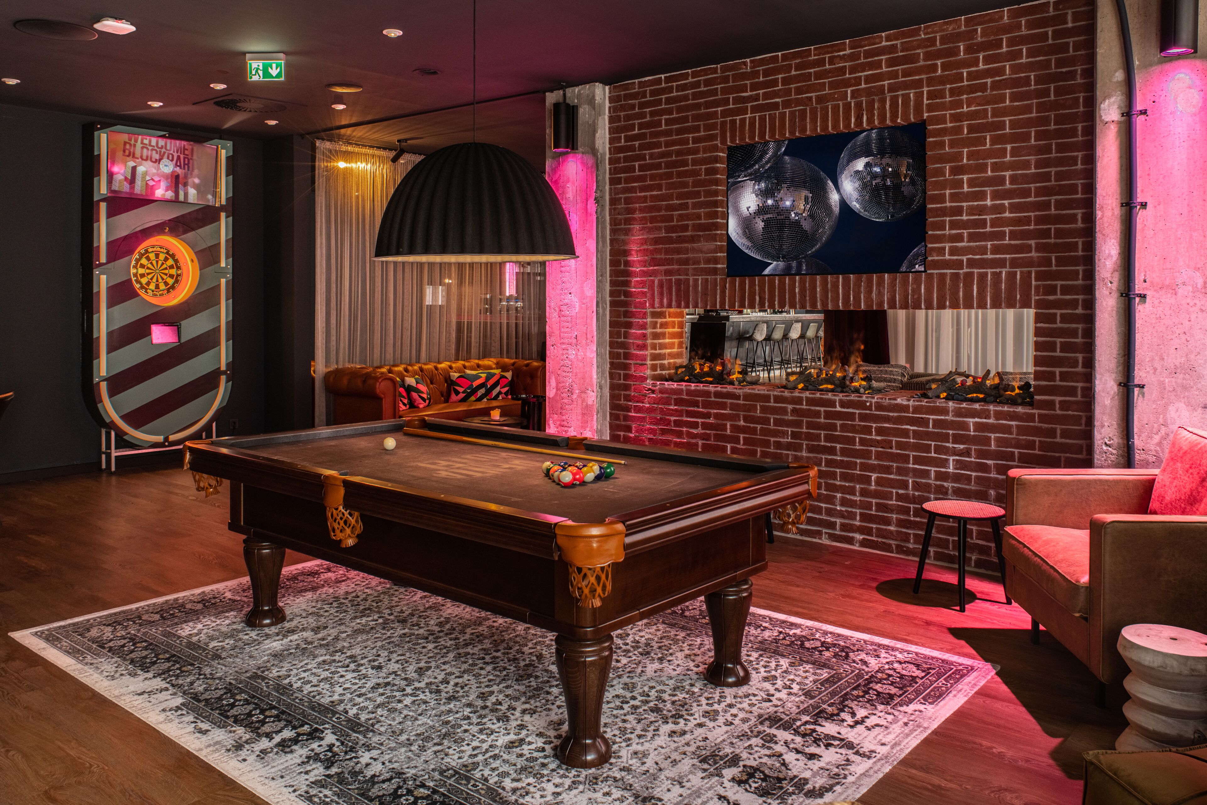 A pool table with balls, a billiard cue, and a dartboard in a room with a brick wall, a bar area, and a cushioned seating area.