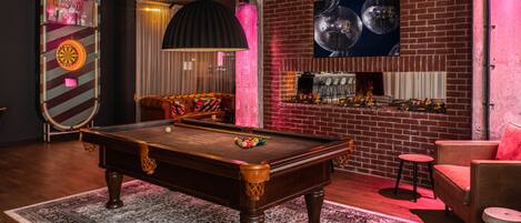 A pool table with balls, a billiard cue, and a dartboard in a room with a brick wall, a bar area, and a cushioned seating area.