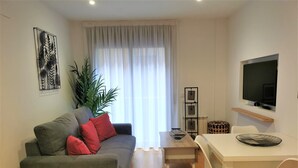 Un salon moderne avec un canapé gris, des coussins rouges, une table basse en bois et une table à manger blanche avec des chaises. Une grande fenêtre avec des rideaux légers laisse entrer la lumière naturelle, et un téléviseur noir est fixé au mur.
