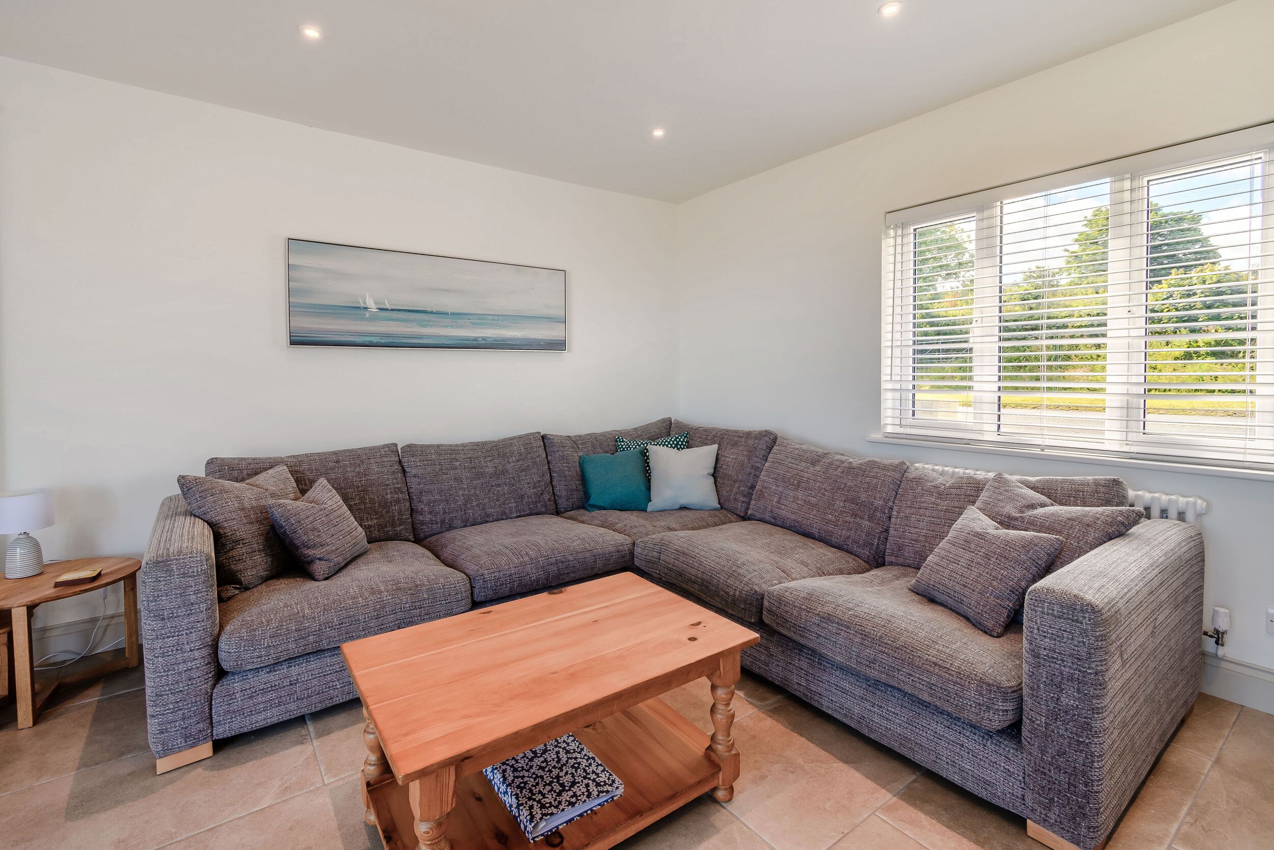 A modern living room with a sectional sofa, a wooden coffee table, a side table, and a window with blinds.