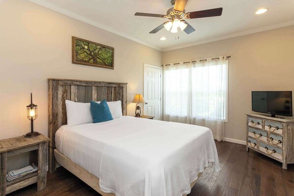 Un dormitorio con cabecera de madera, cama con ropa de cama blanca y almohada azul, mesita de noche con lámpara, televisor sobre un tocador y ventana con cortinas.