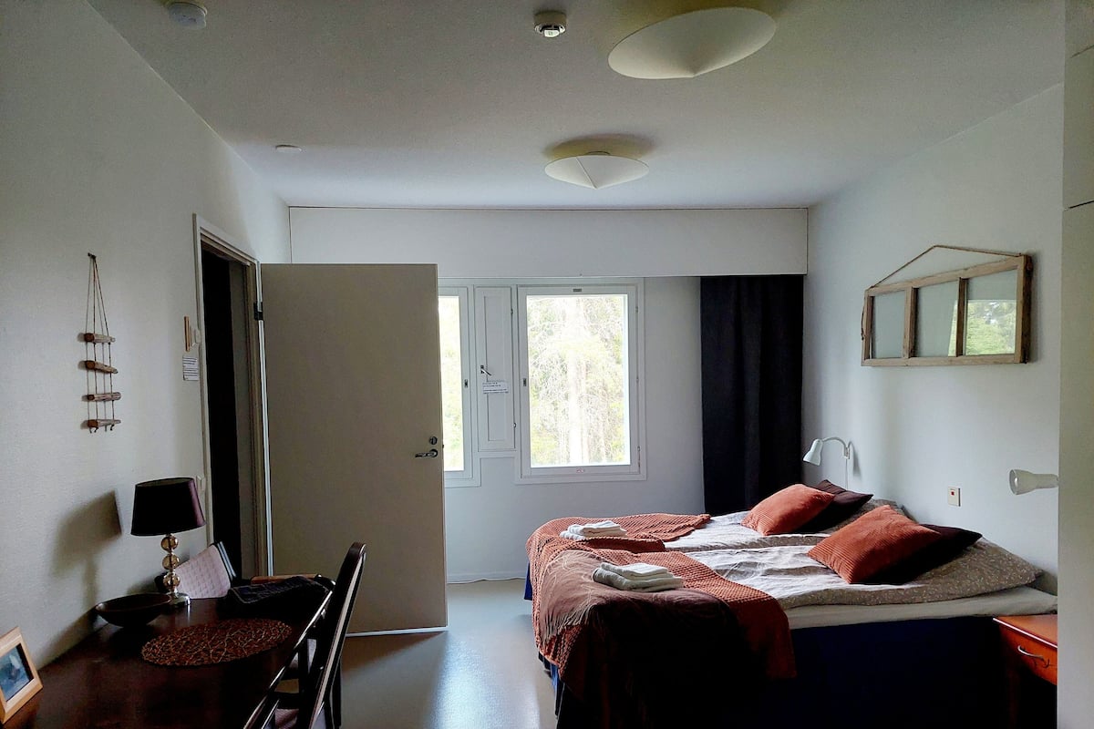 A bedroom with a bed, bedside table, lamp, desk, chair, and a window with curtains.