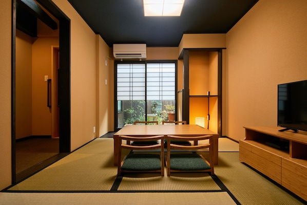 A room with tatami mats, a wooden table with chairs, a TV, and a window with a view of greenery.