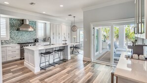 A modern kitchen with white cabinetry, a large island with bar stools, and a view to a patio area. - Sunset Point - Dreamy Beach House, Hot Tub, Roof Top Deck, Great Location! (Treasure Island)