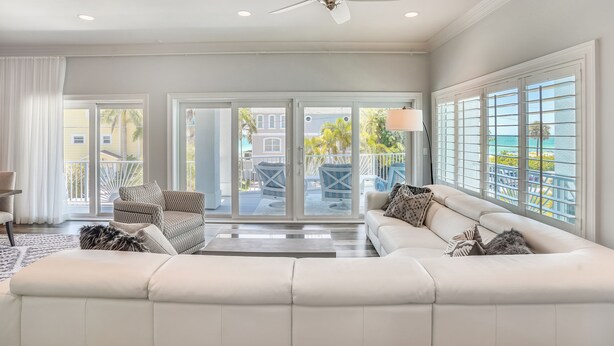 A spacious living room with a large white sofa, a ceiling fan, and a view of a balcony with outdoor furniture. - Sunset Point - Dreamy Beach House, Hot Tub, Roof Top Deck, Great Location! (Treasure Island)