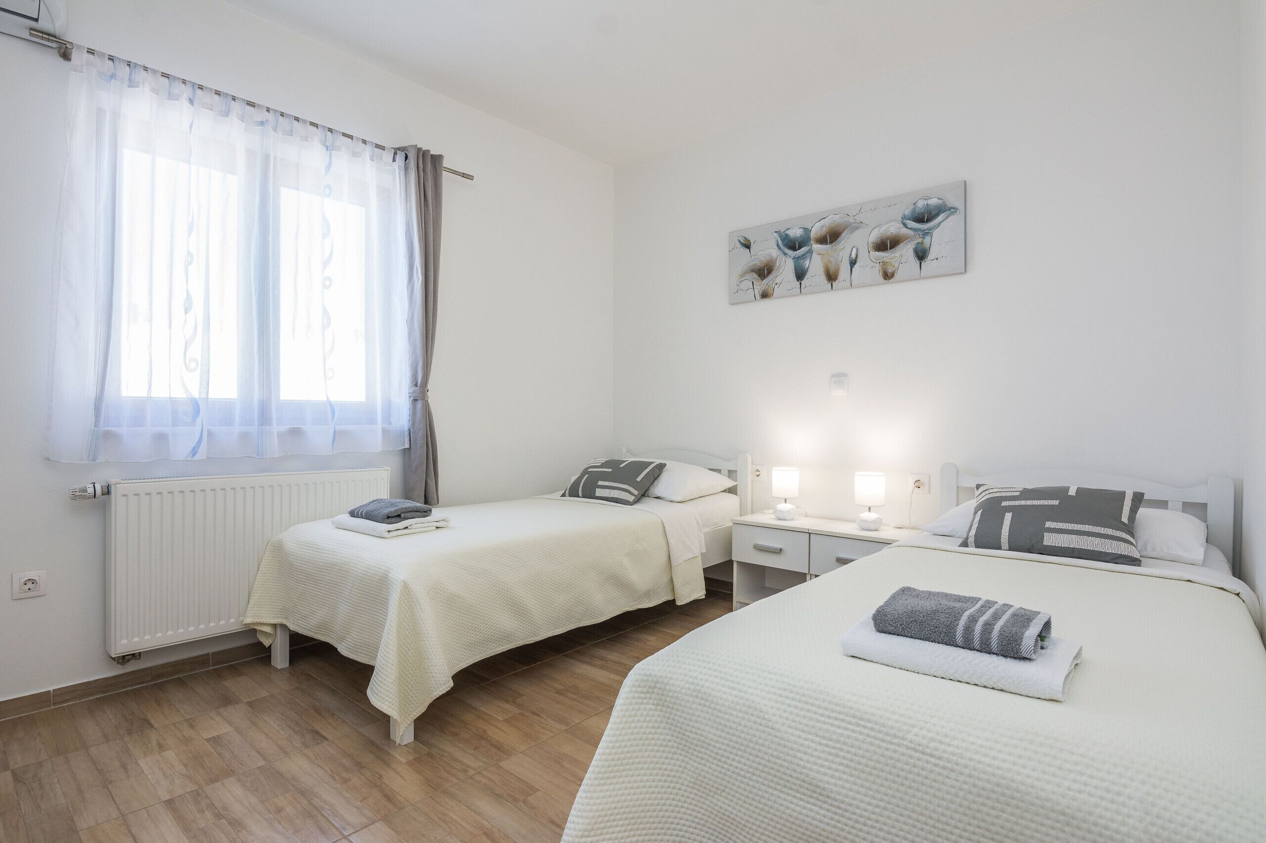 Une chambre avec deux lits, une table de chevet, une lampe fixée au mur et une fenêtre avec des rideaux légers.