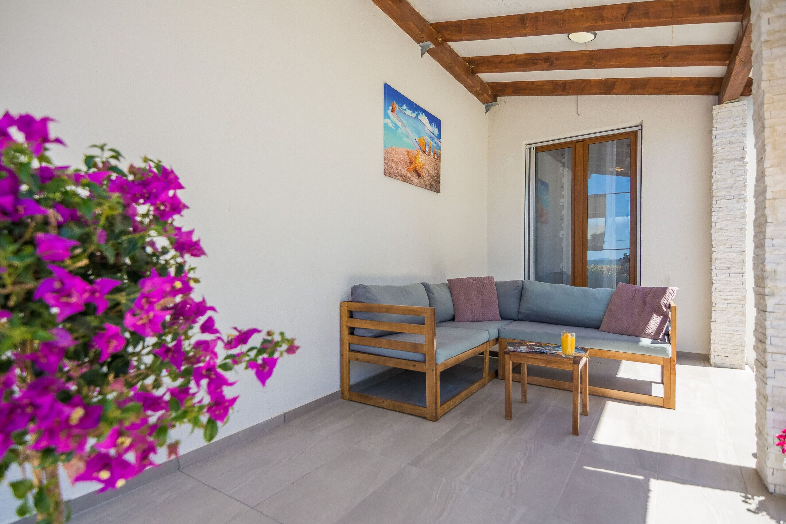 A sunlit patio with a wooden sofa and coffee table, a potted plant, and a painting on the wall.