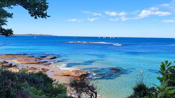 Un paysage côtier avec une eau d’un bleu limpide, une côte rocheuse et des voiliers qui se dessinent à l’horizon.
