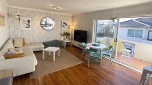 Un salon moderne avec un canapé blanc, une petite table blanche et une table basse ronde. On y trouve un téléviseur posé sur un meuble blanc, une plante en pot et un balcon avec des chaises jaunes.