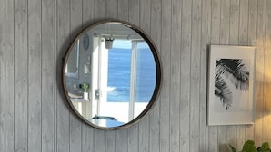 Un miroir rond fixé sur un mur en bois, qui reflète une porte et une vue sur l’océan.