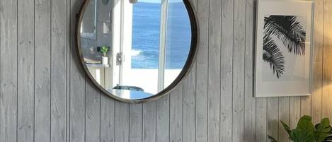 Espejo redondo sobre una pared de madera, que refleja una puerta y vistas al océano.