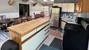 A modern kitchen with a central island, a dining area with a wooden table, and a living room with a fireplace. - Cabin in the woods (Wolverine)