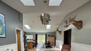 A spacious living room with a chandelier, a fireplace, mounted deer heads, and a dining area in the background. - Cabin in the woods (Wolverine)