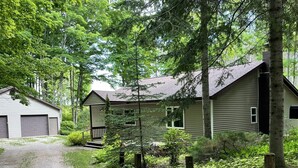 A two-story house with a porch, surrounded by dense greenery and trees. - Cabin in the woods (Wolverine)