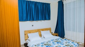 A bedroom with a wooden headboard, a floral bedspread, and blue curtains. - Green Eskapes Suites (Kigali)